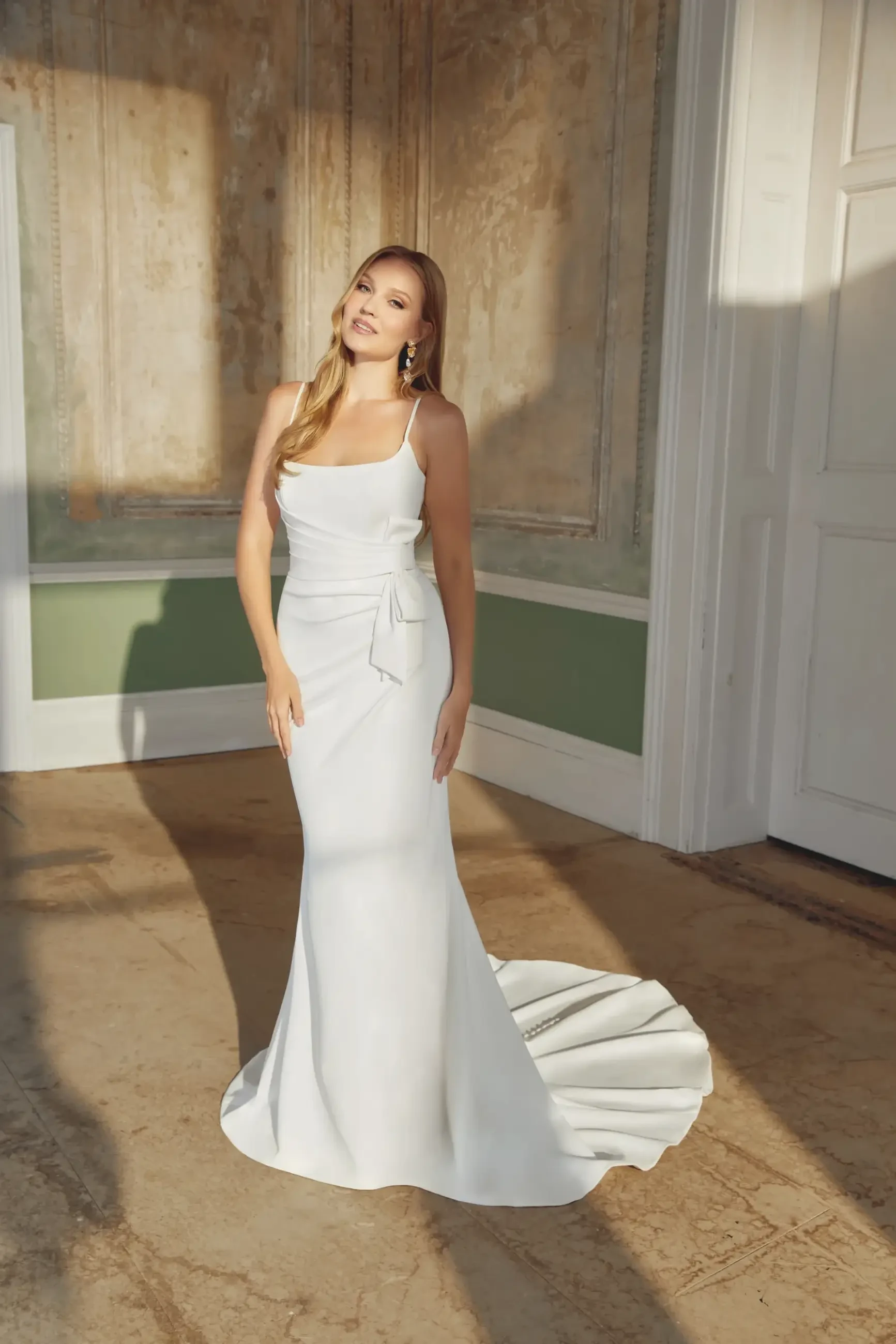 Woman in floor-length, sleek white gown with a bow, standing in a sunlit room with textured walls and shadow patterns, exuding elegance and grace.