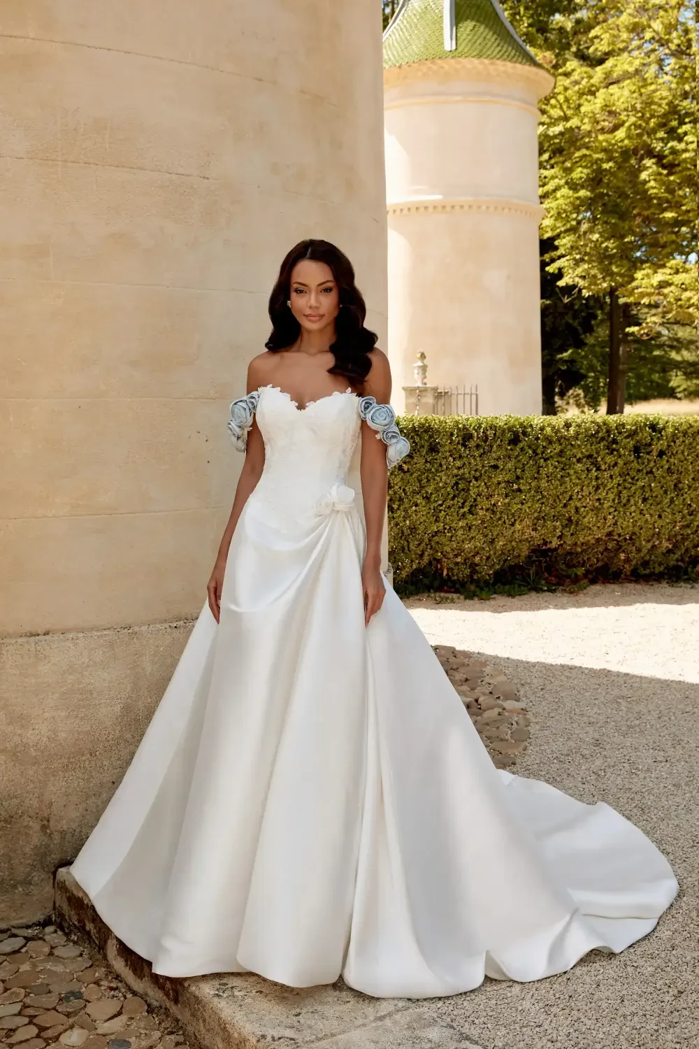 A woman in an elegant off-shoulder wedding dress stands in front of a stone wall, surrounded by greenery.