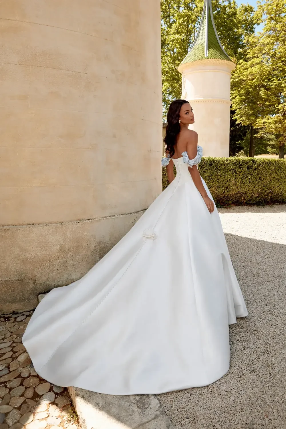 A woman in an elegant off-shoulder wedding dress stands in front of a stone wall, surrounded by greenery.
