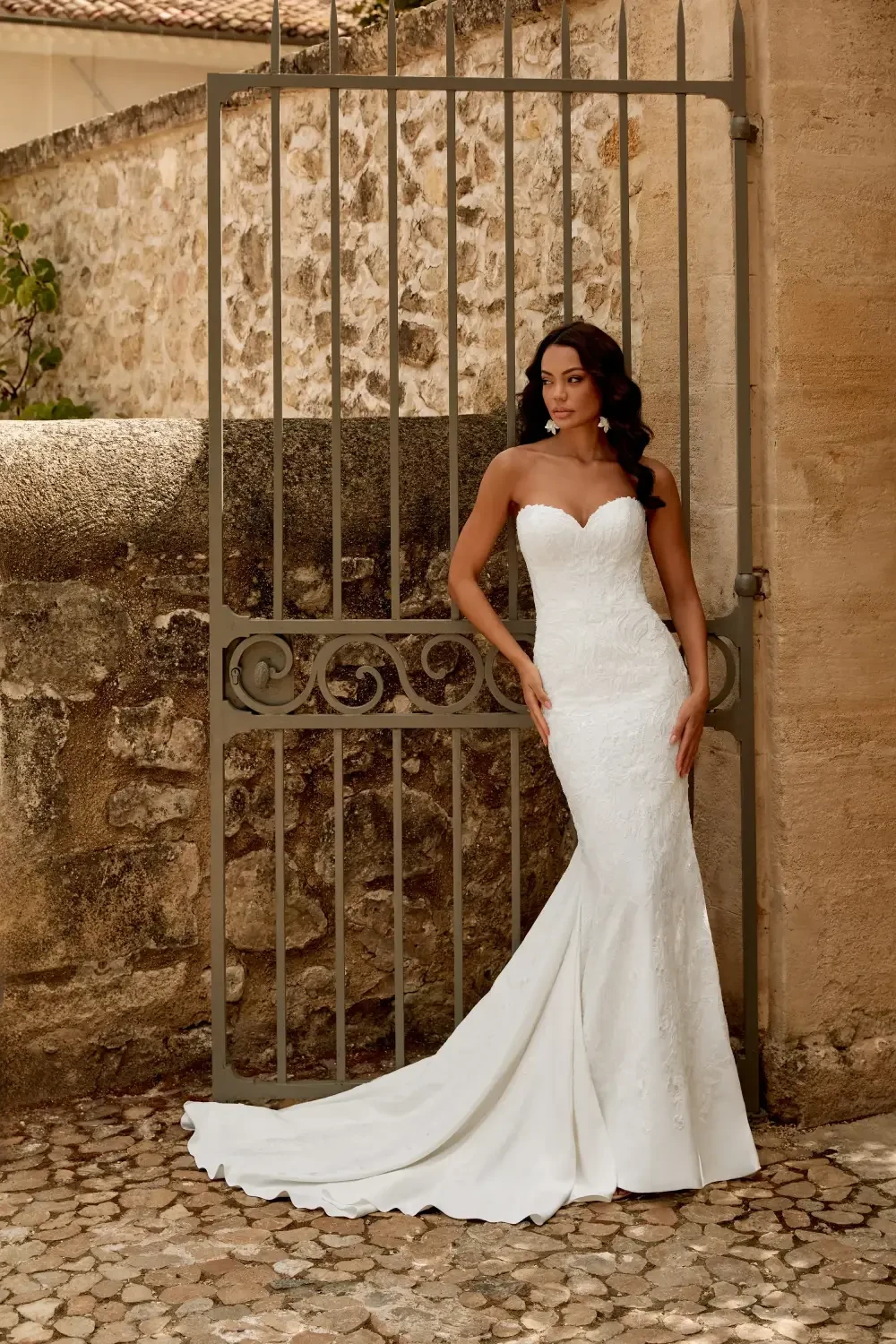 A woman in a form-fitting white wedding dress stands gracefully by a wrought iron gate, set against a textured stone wall.