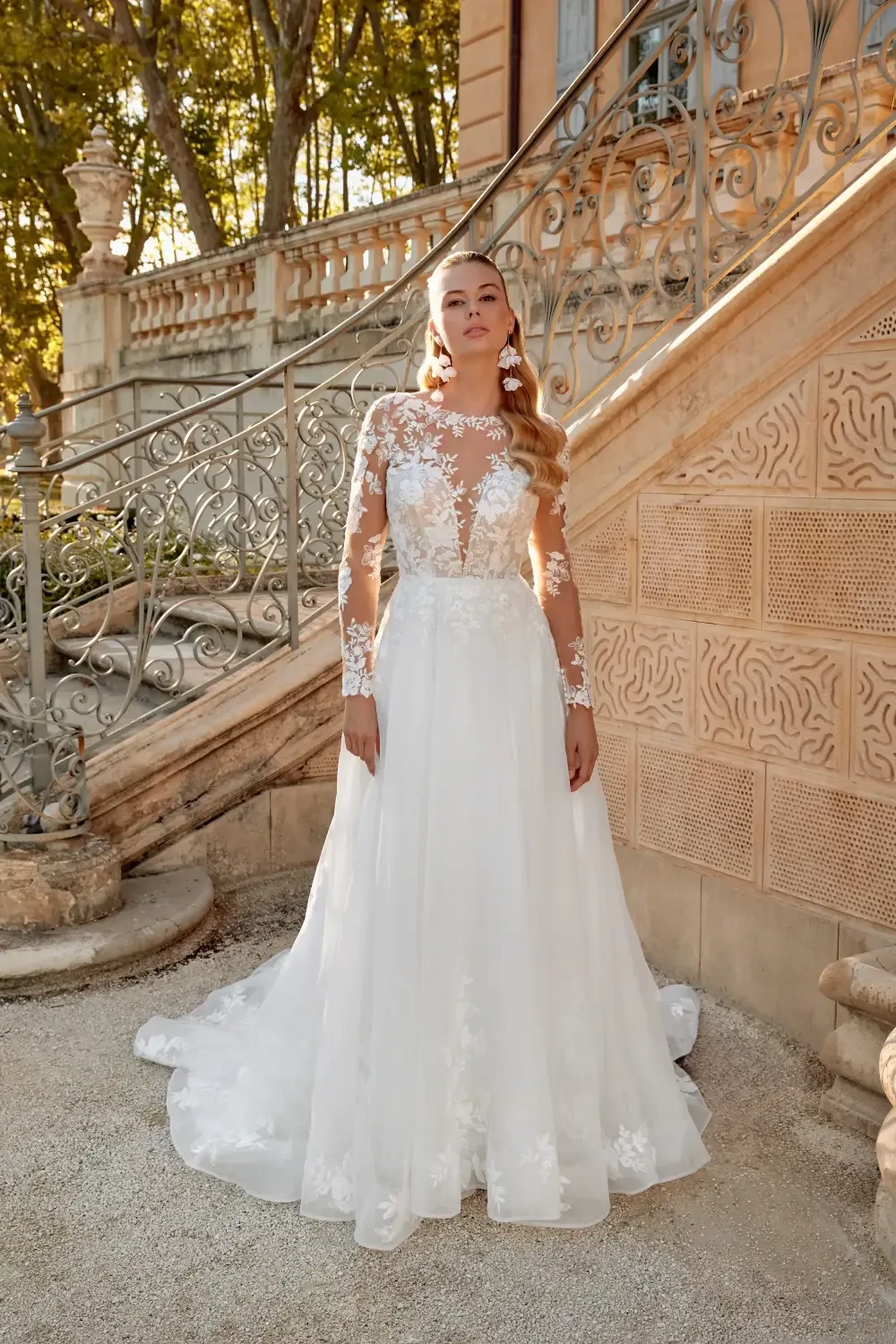 A woman in a white wedding dress with long sleeves stands on a staircase, surrounded by greenery and ornate architecture.