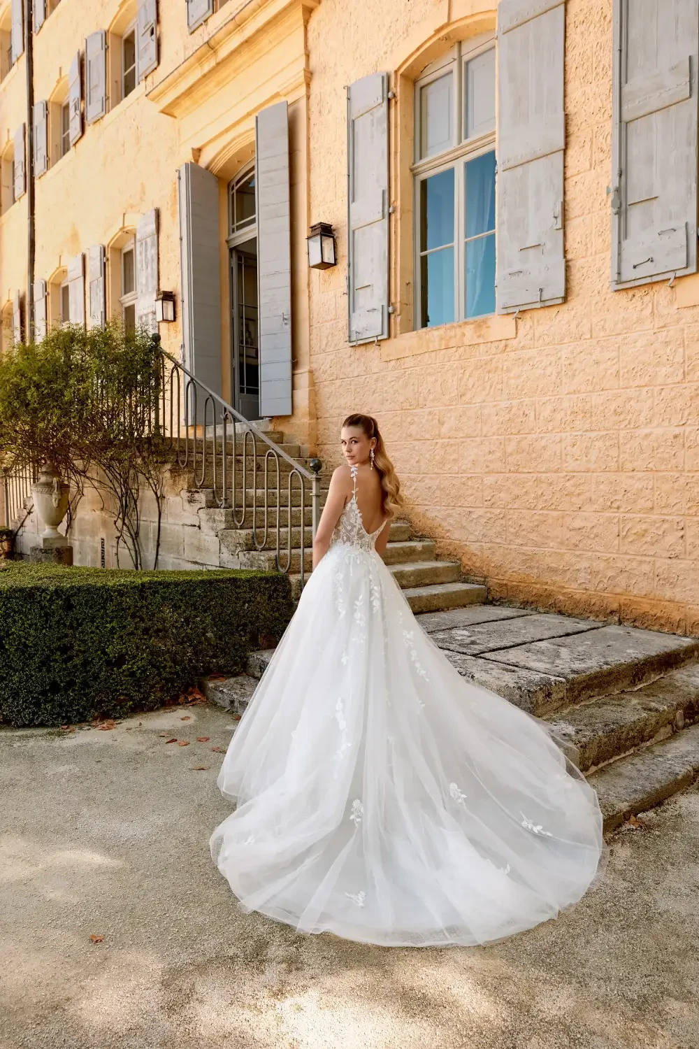 A bride in a stunning white wedding dress stands outdoors in front of a picturesque building with large windows and stone steps.