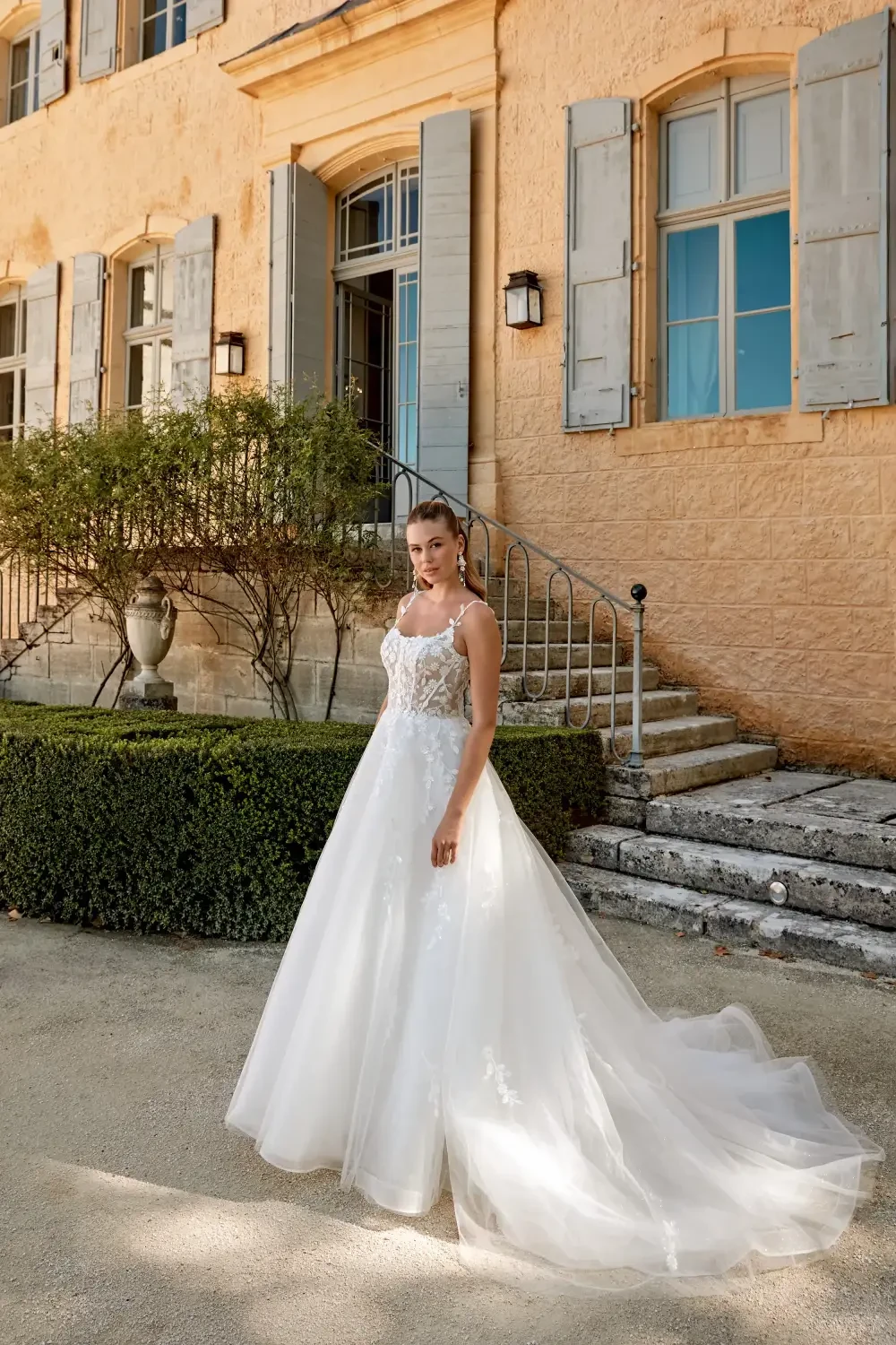 A bride in a stunning white wedding dress stands outdoors in front of a picturesque building with large windows and stone steps.