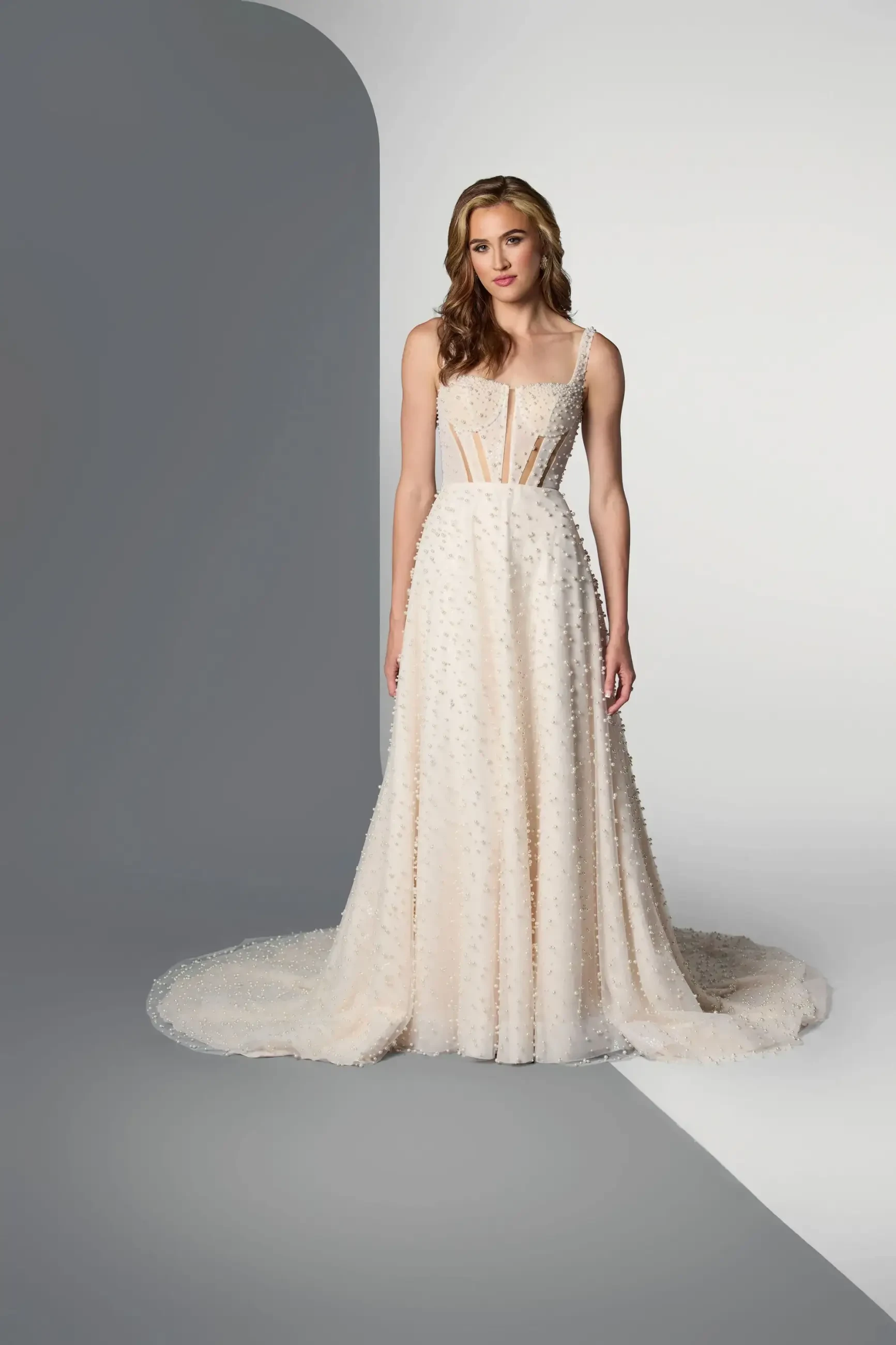 Model is wearing a sleeveless A-line nude-champagne wedding dress with 3D pearl beadwork, illusion cutouts, and corset bodice.