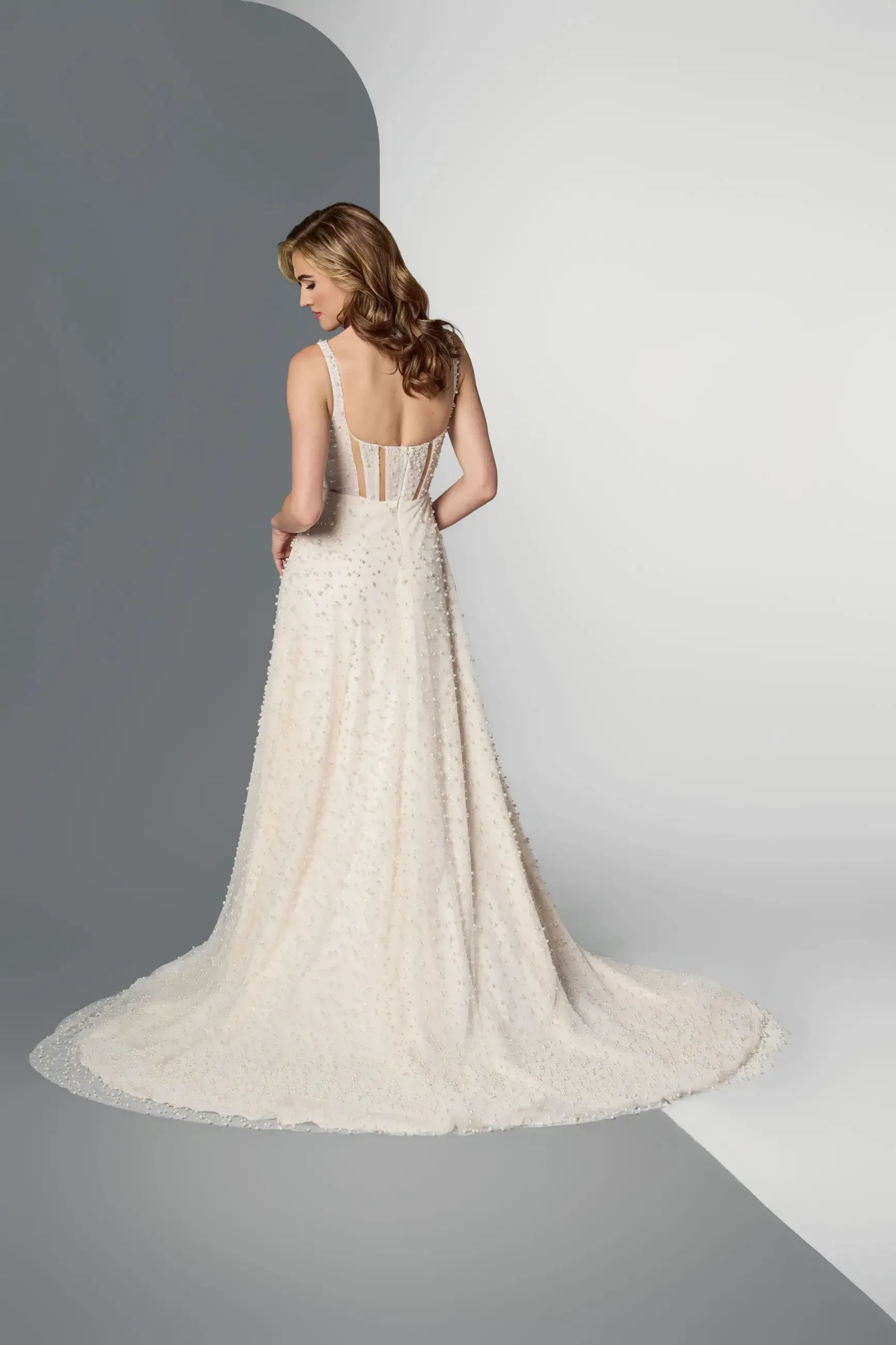 Model is wearing a sleeveless A-line nude-champagne wedding dress with 3D pearl beadwork, illusion cutouts, and corset bodice.