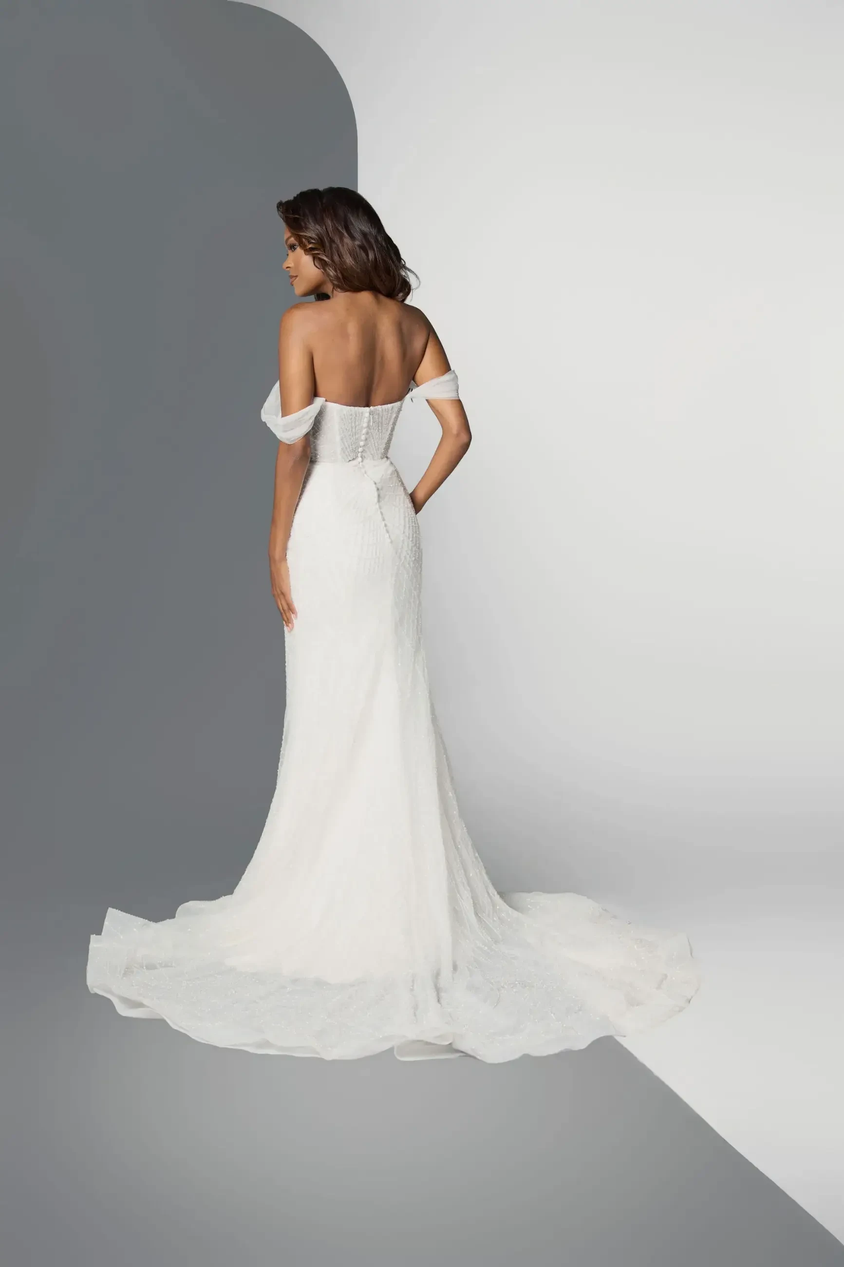 Model is wearing an off-the-shoulder fit-and-flare wedding dress with a corset bodice and train.