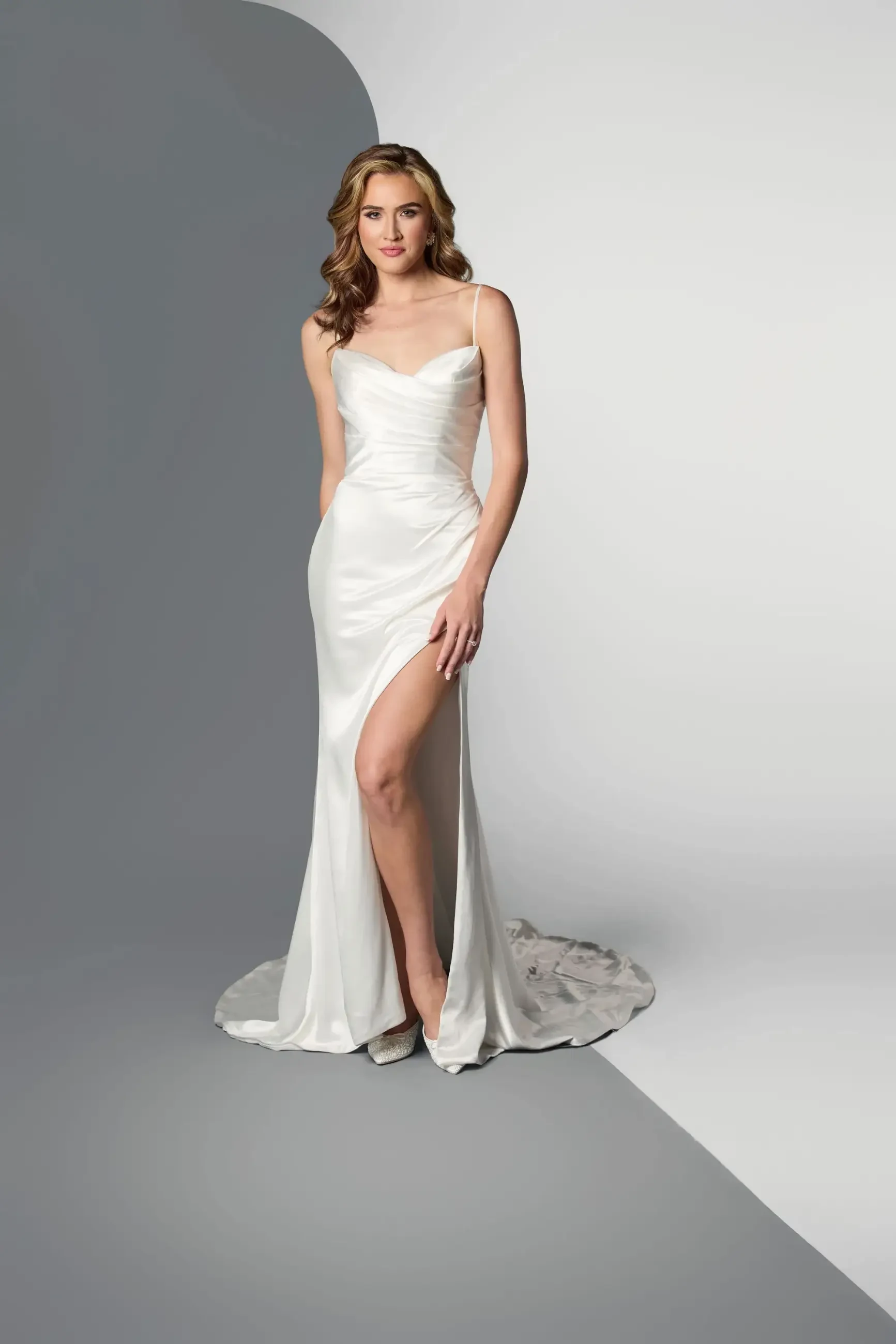 Model is wearing a sleeveless sheath wedding dress with spaghetti straps, a slit, and a silky minimalist design.