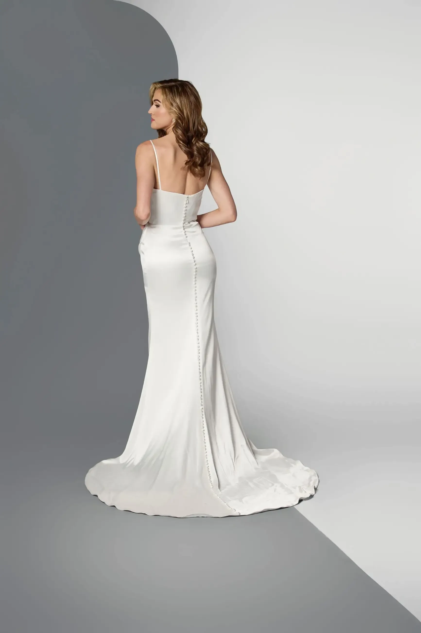 Model is wearing a sleeveless sheath wedding dress with spaghetti straps, a slit, and a silky minimalist design.