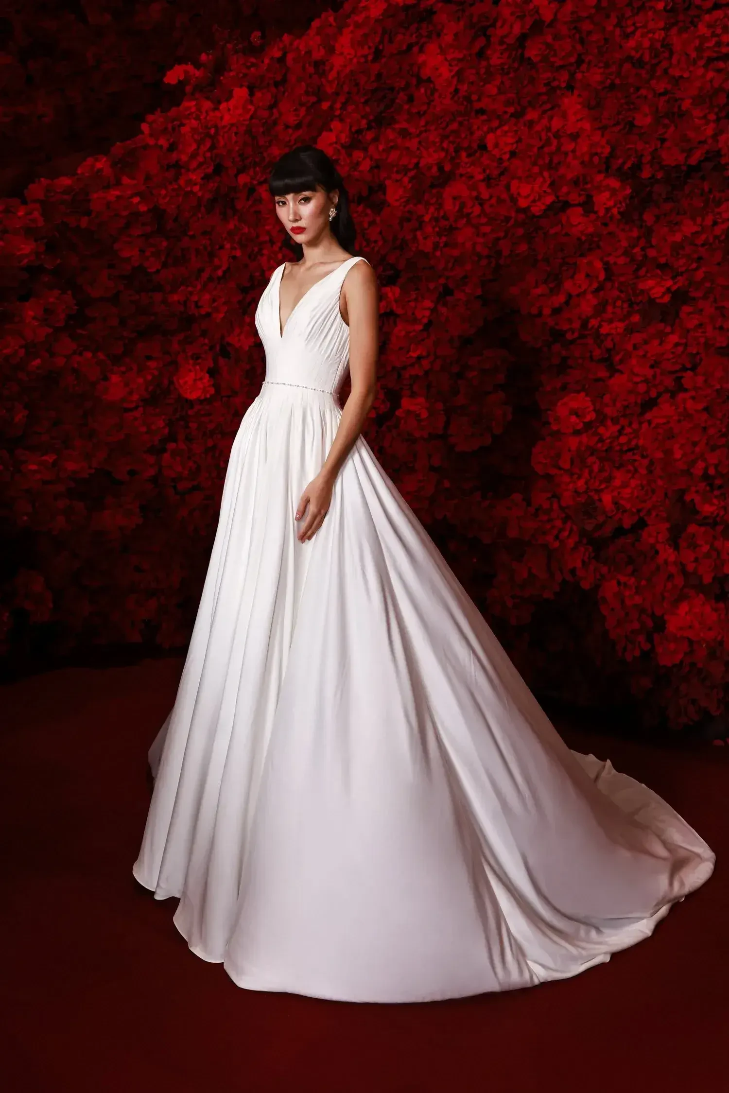 A woman in an elegant white gown stands against a vibrant red floral wall. The scene exudes sophistication and contrast with dramatic tones.