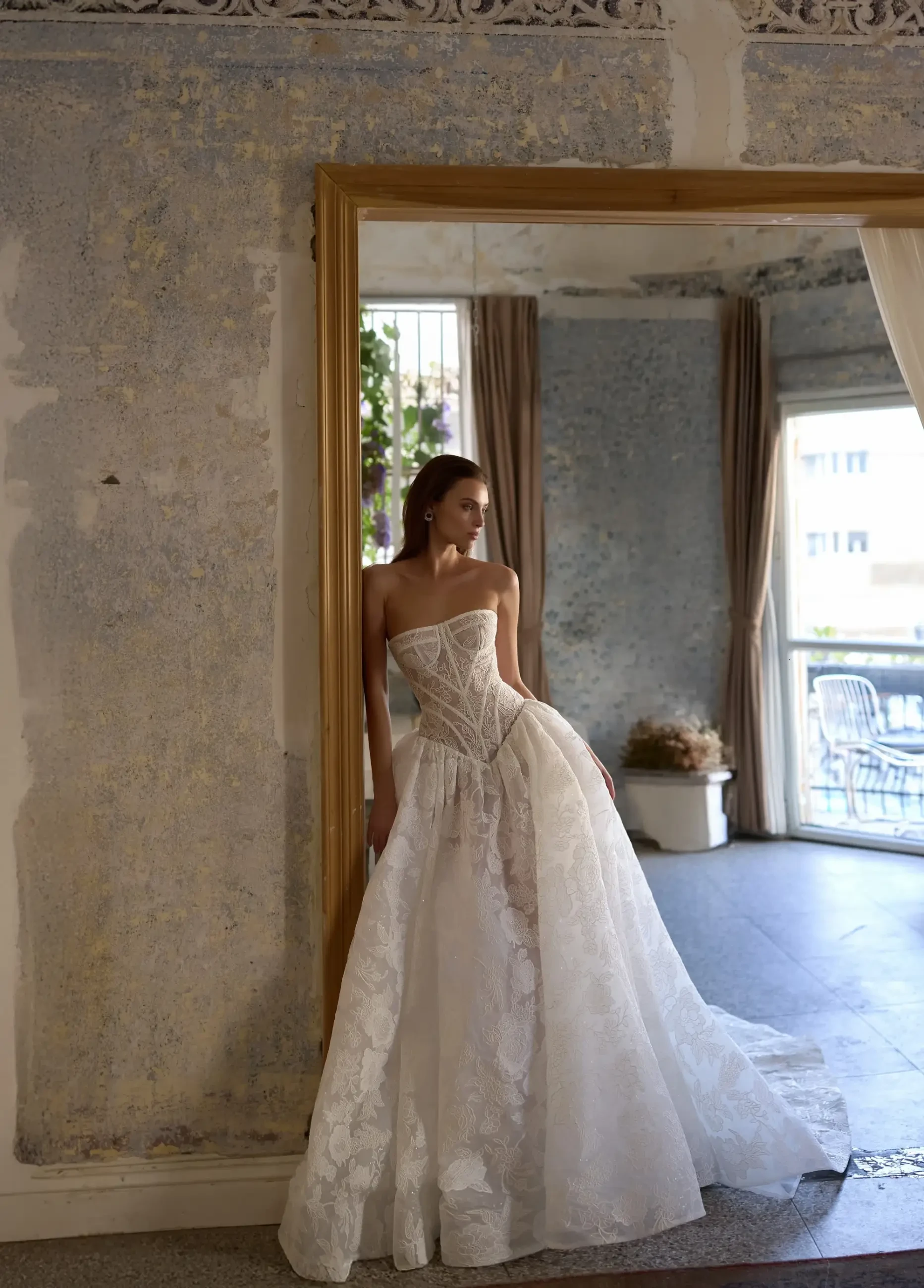 A woman in a strapless, intricate lace wedding gown stands by a rustic, textured wall in a softly lit room. The mood is elegant and serene.