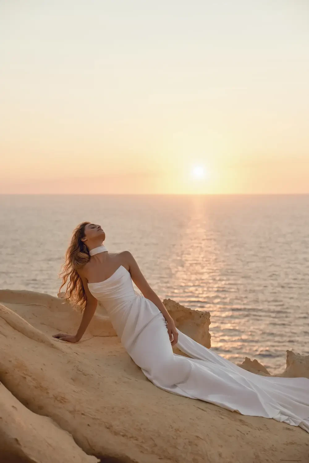 A woman in a flowing white dress poses on a rocky ledge by the sea during sunset, with the sun setting over the horizon.