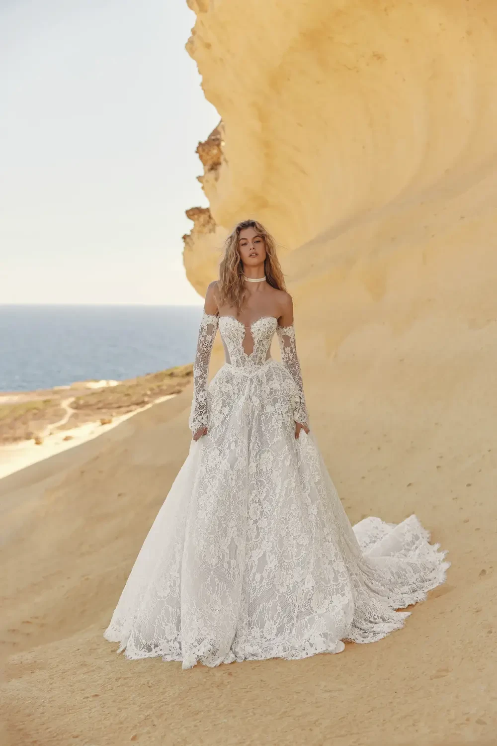 A model wearing a stunning white lace wedding gown with off-the-shoulder sleeves standing against a sandy cliff by the sea.