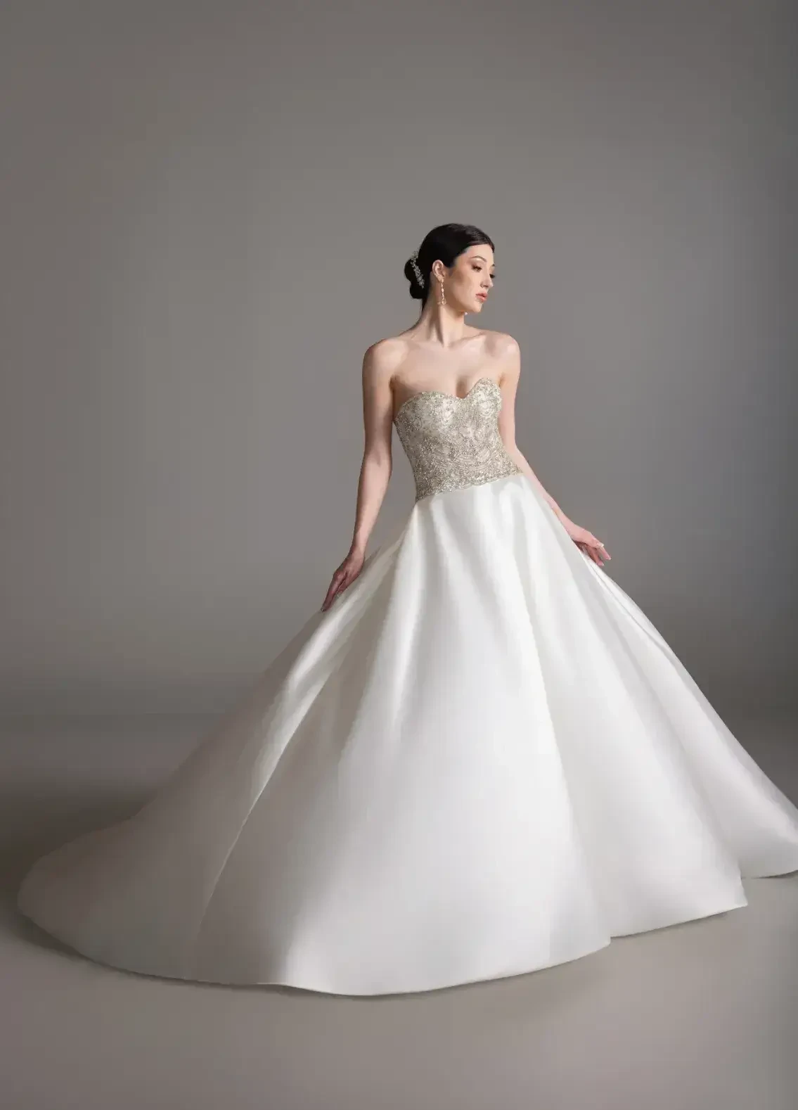 A woman in a strapless white gown with intricate beading stands elegantly against a neutral background.