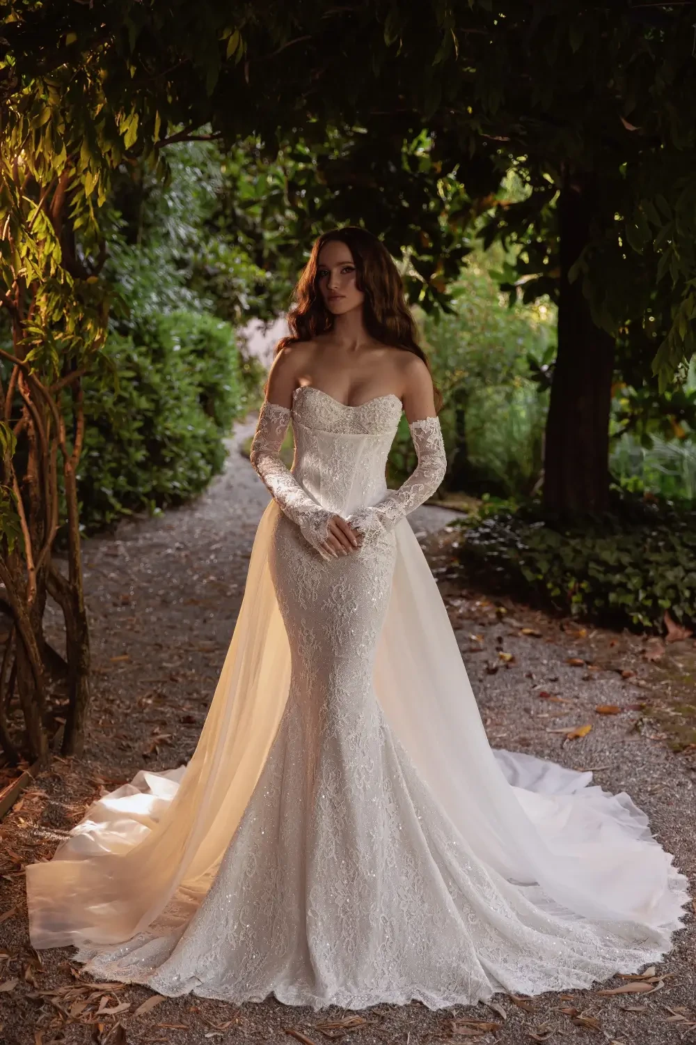 A woman wearing an elegant wedding dress with long lace sleeves, standing in a lush, green garden pathway.