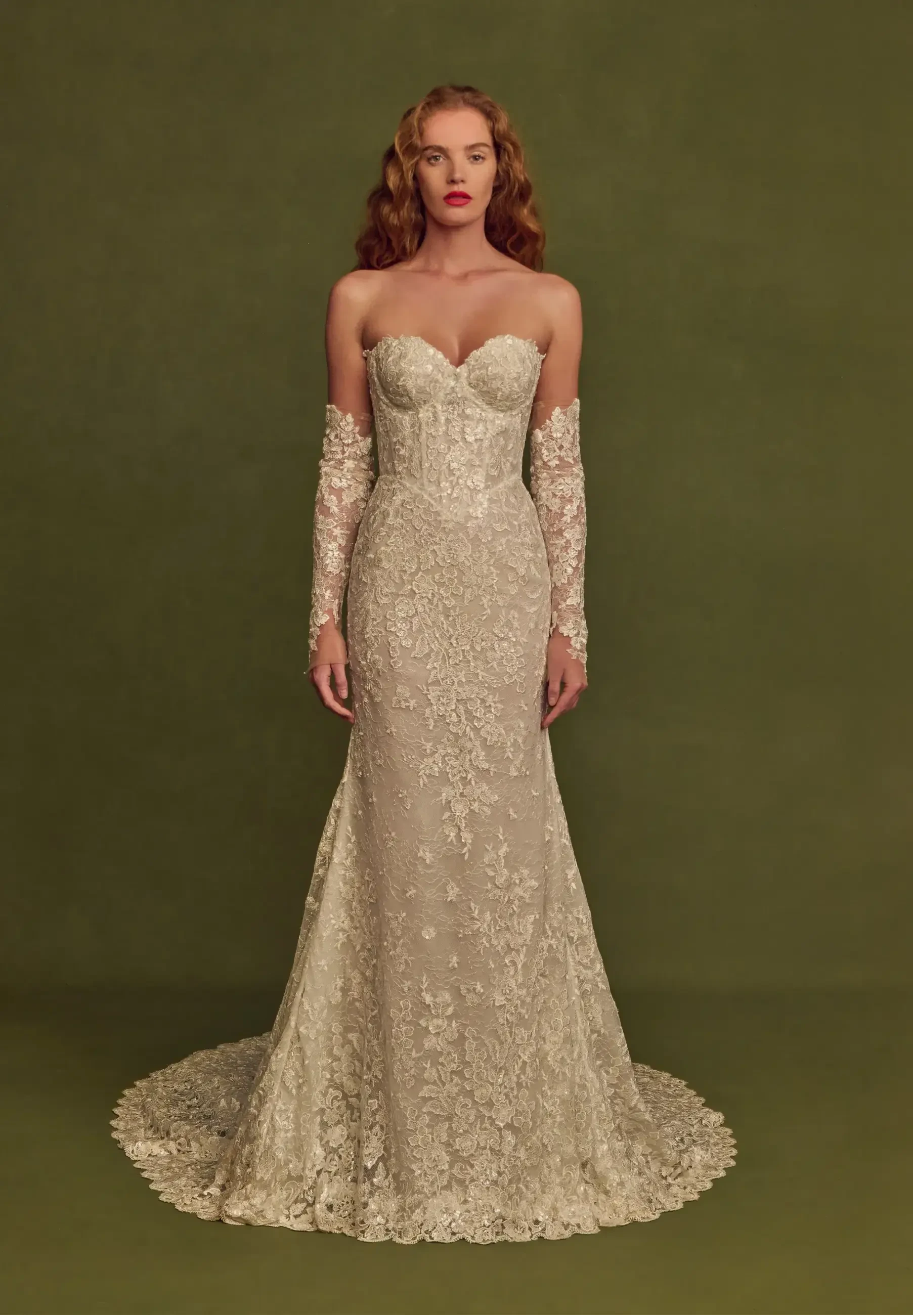 A woman in a strapless lace wedding gown with long lace sleeves and a fitted silhouette, standing against a green background.