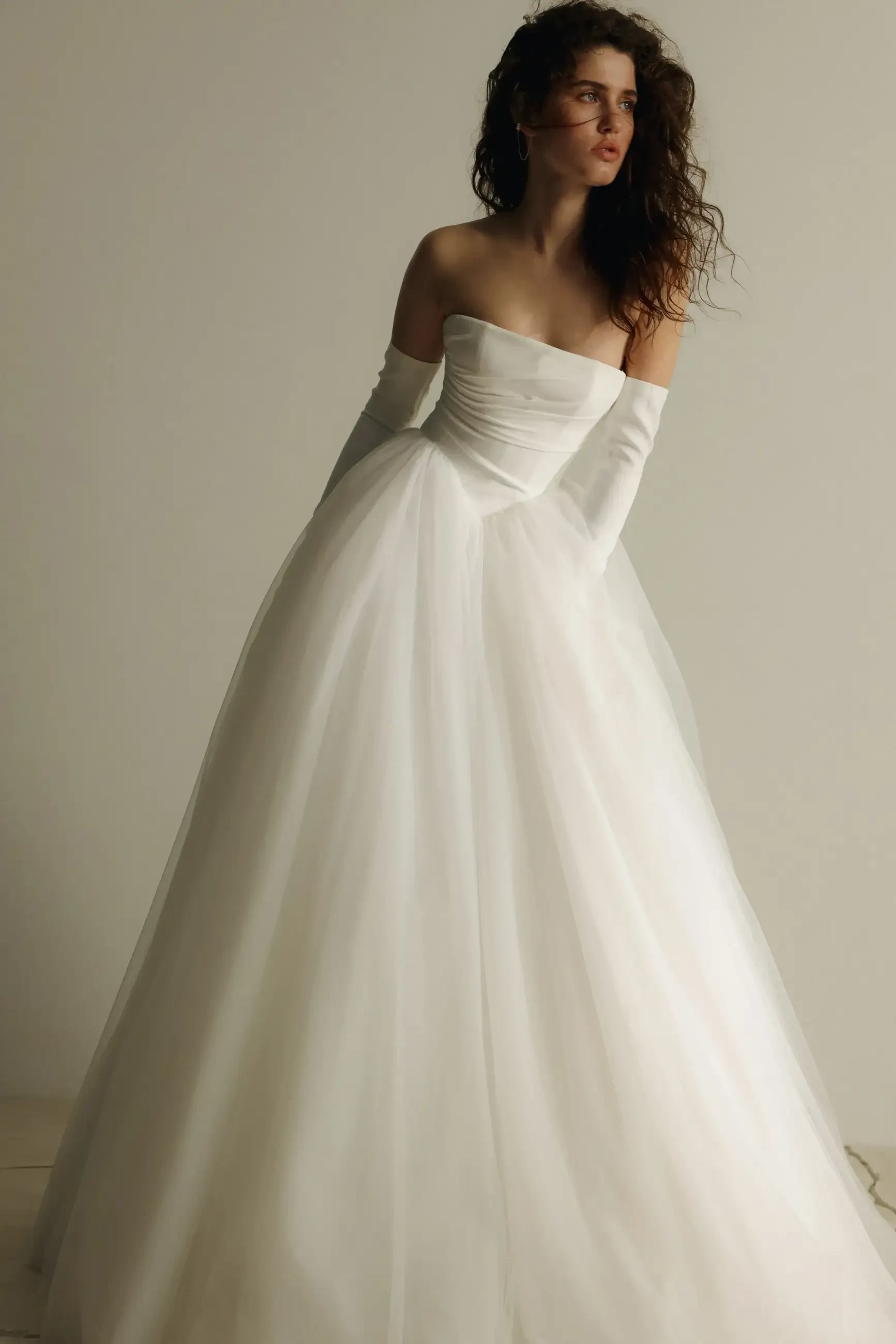 A woman in a strapless white ball gown with long gloves stands against a plain backdrop. Her wavy hair and serene expression evoke elegance.