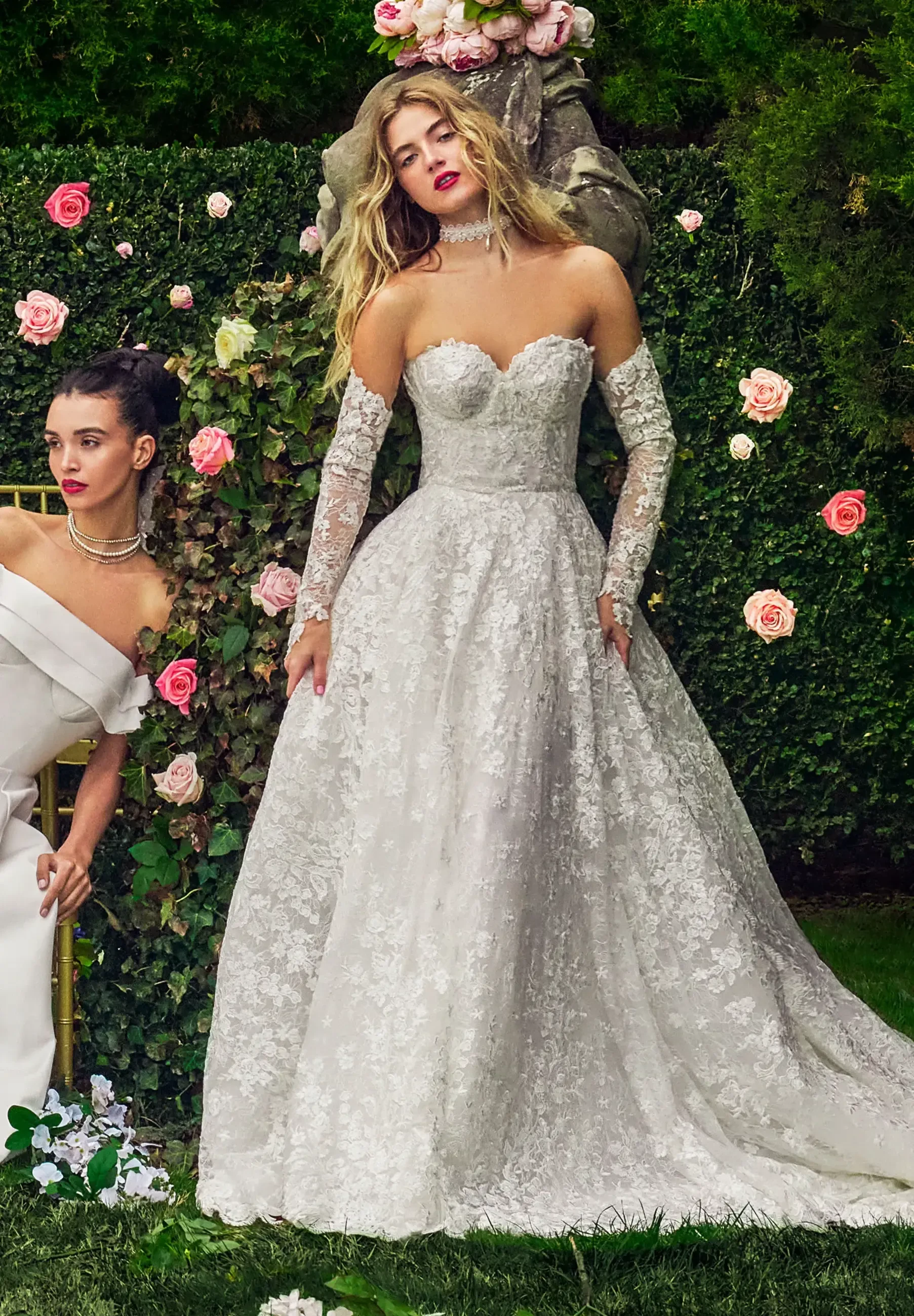 Two women in elegant dresses posing in a garden adorned with roses. One woman is wearing a strapless white gown, while the other is in a long-sleeved, off-the-shoulder white dress.