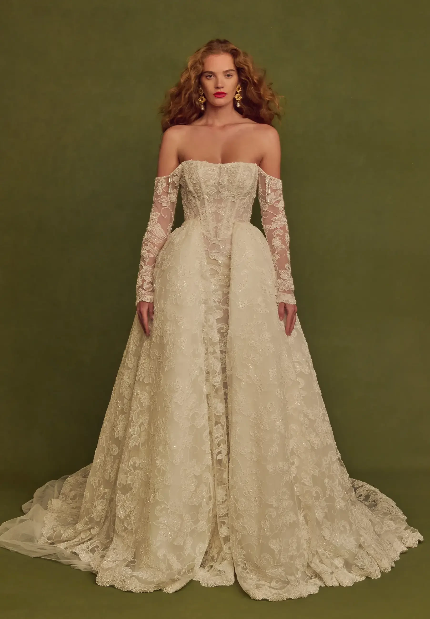 A woman in a strapless, ivory wedding gown with intricate lace detailing and long lace gloves, standing against a green background.