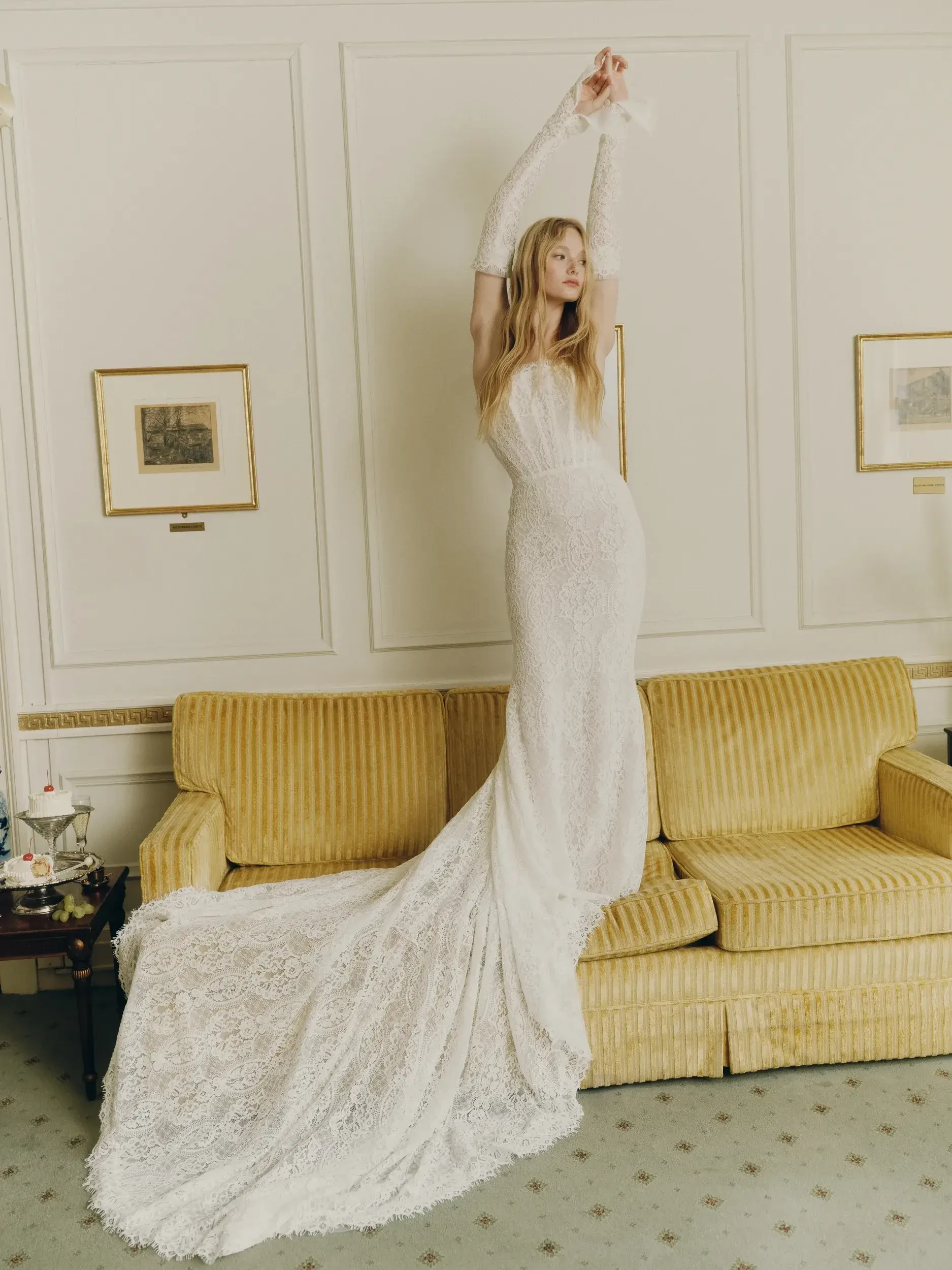 A model in a long, elegant white lace gown poses with arms raised on a yellow couch in a sophisticated room, conveying grace and sophistication.