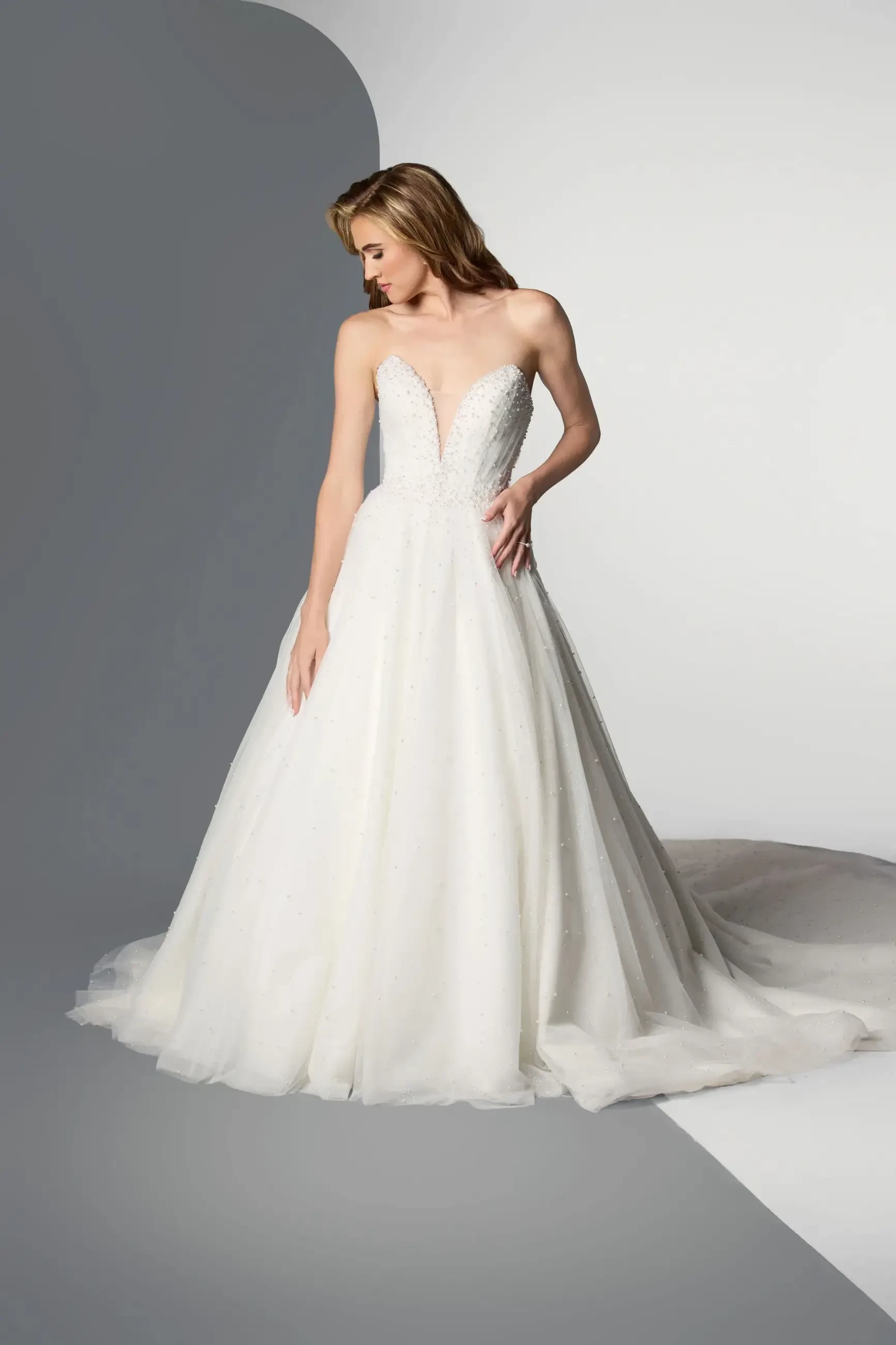 A woman in an elegant, strapless white wedding gown with a fitted bodice and flowing skirt stands against a gray and white background, exuding grace.