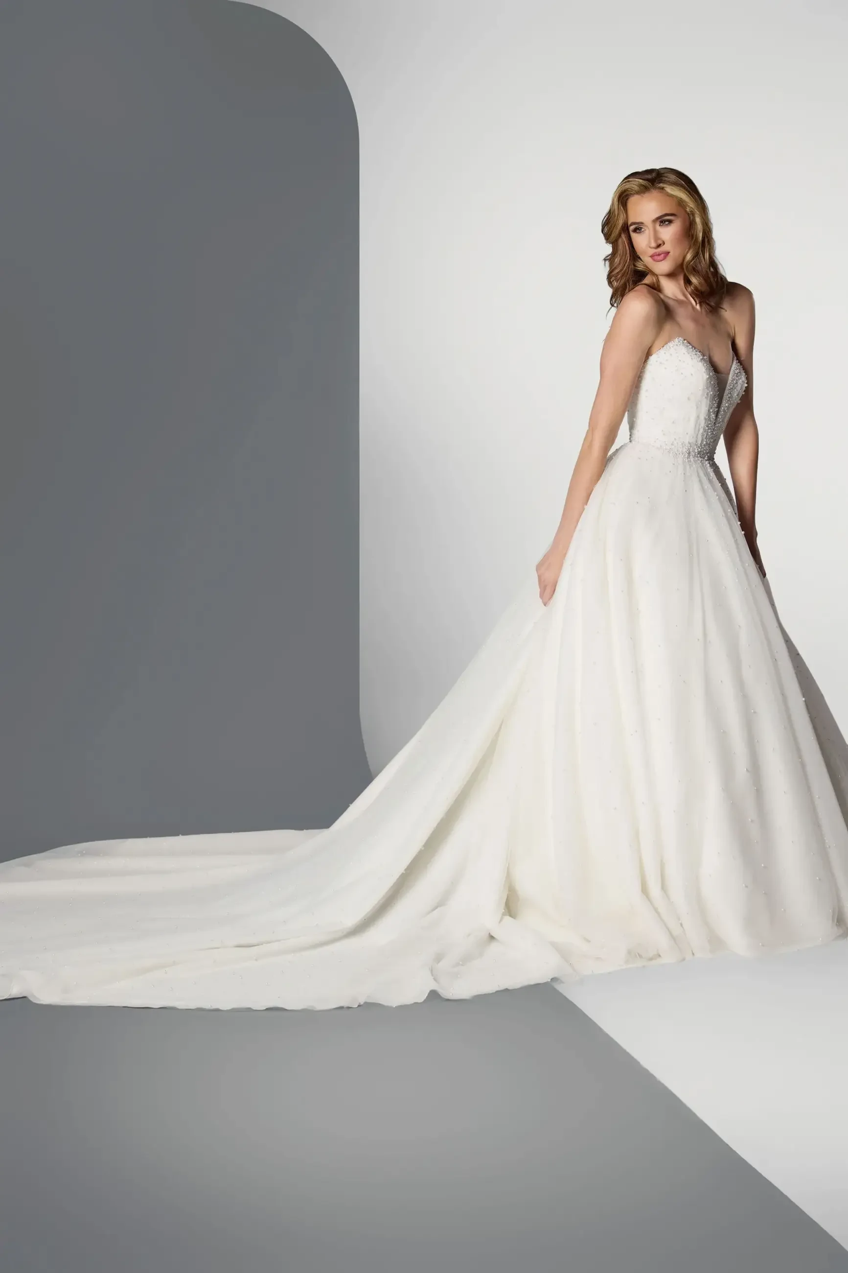 A model wearing a white wedding dress poses against a minimalist background.