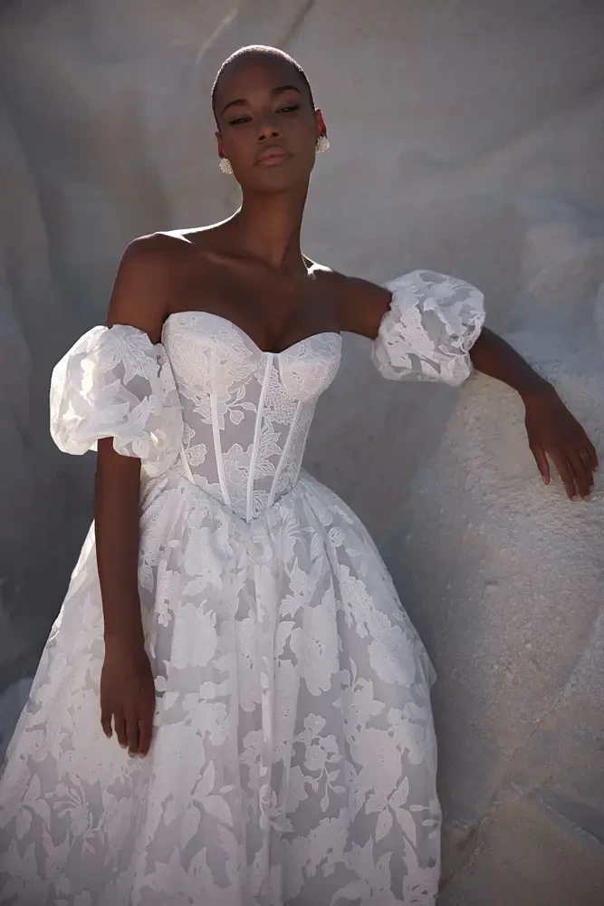 Model is wearing an off the shoulder wedding dress with a basque waist and silky floral details