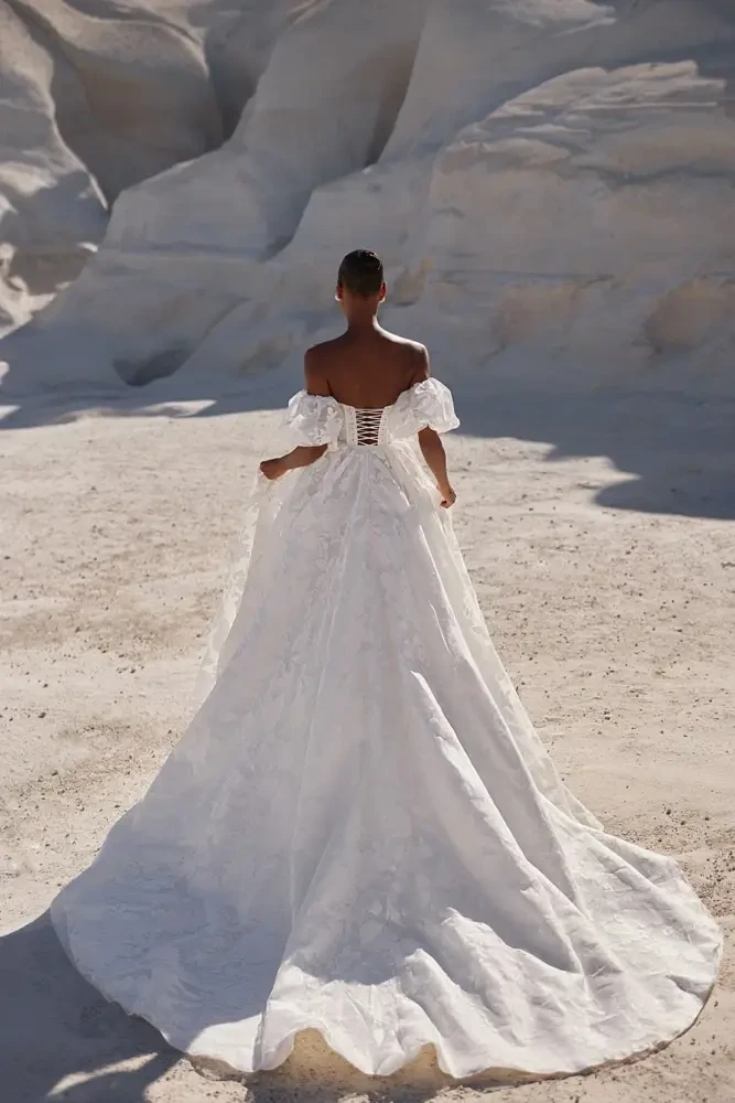 Model is wearing an off the shoulder wedding dress with a basque waist and silky floral details