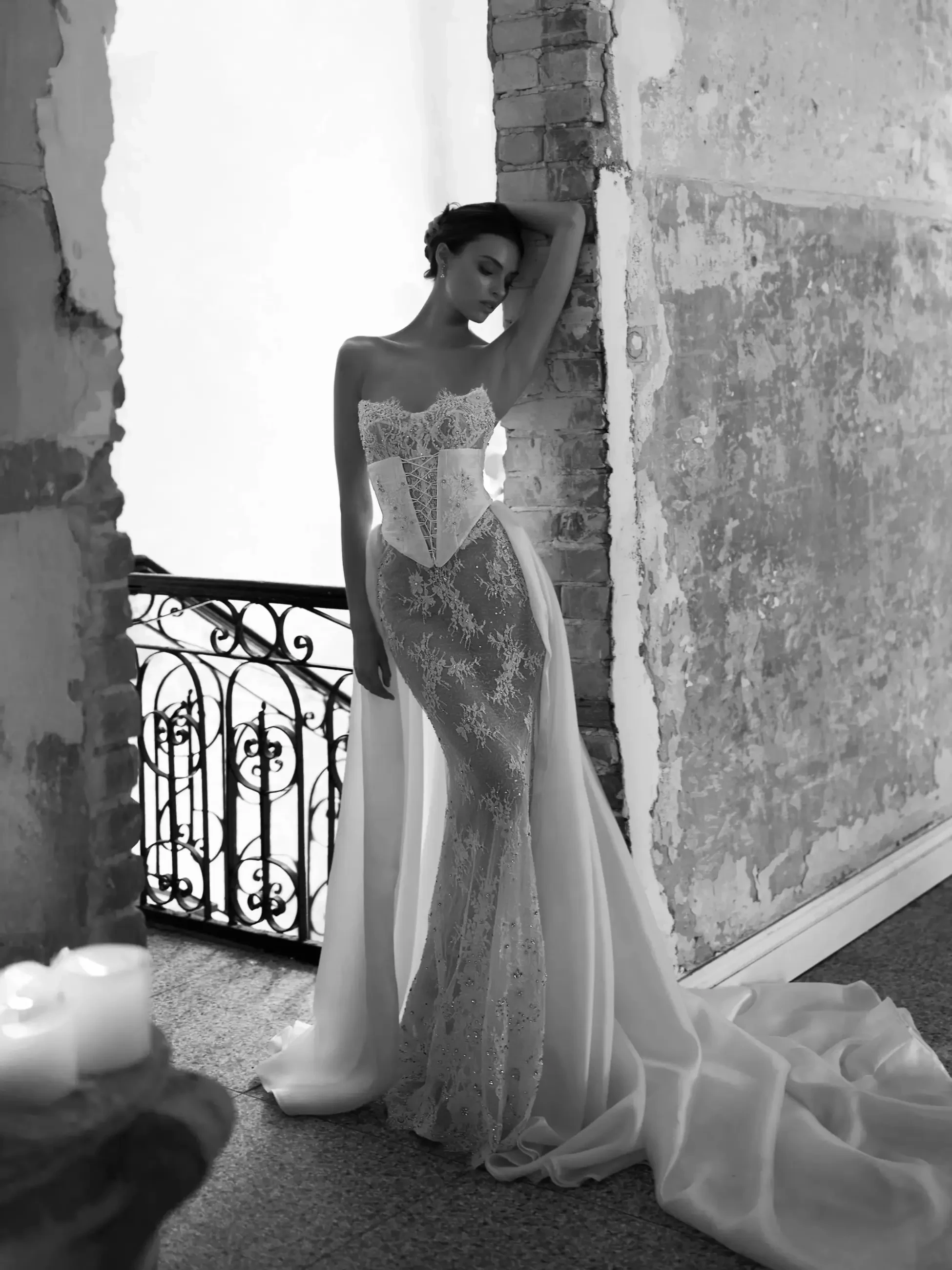 A woman in a stunning, detailed white gown stands gracefully against a textured wall, with elegant candles nearby.