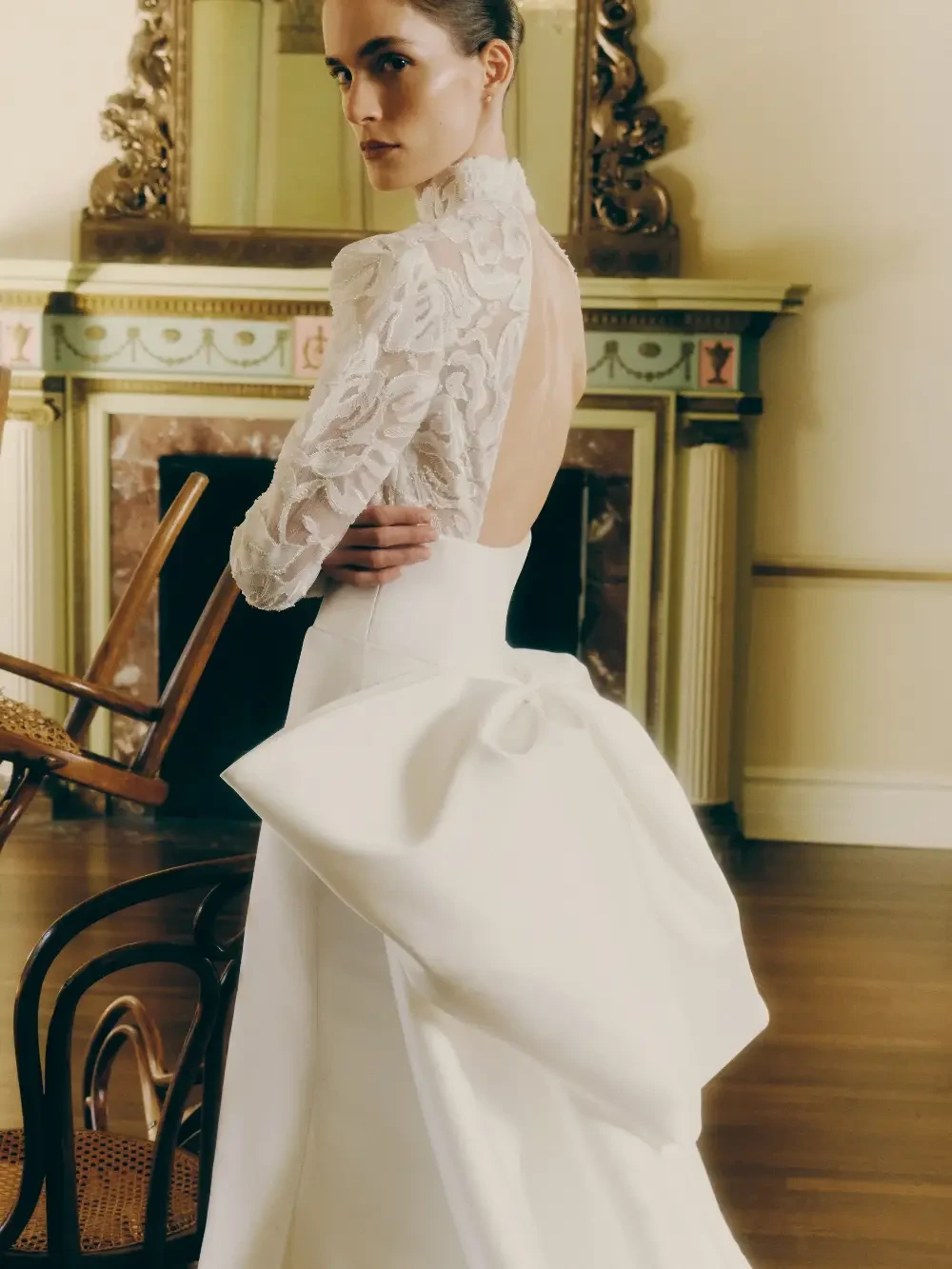A woman in a wedding dress with a high neckline and an open back, featuring a large bow at the waist, standing in a room with elegant decor.