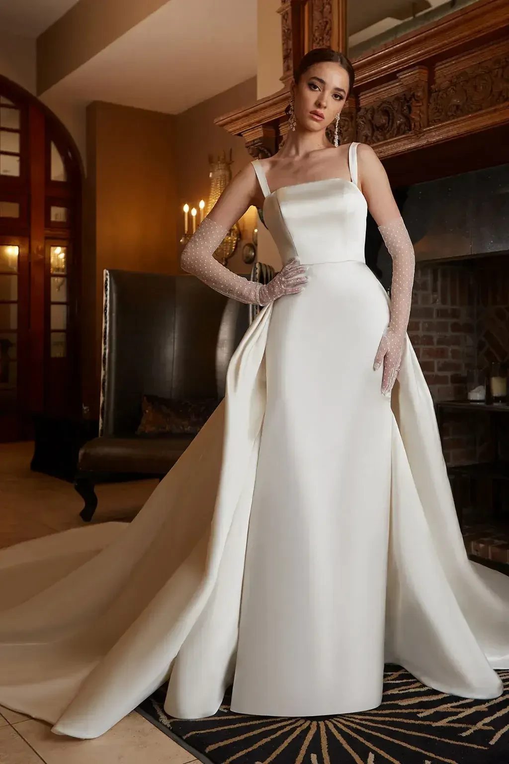 A model wearing an elegant, white satin wedding gown with a fitted bodice and a flowing train, posed in an upscale interior setting.