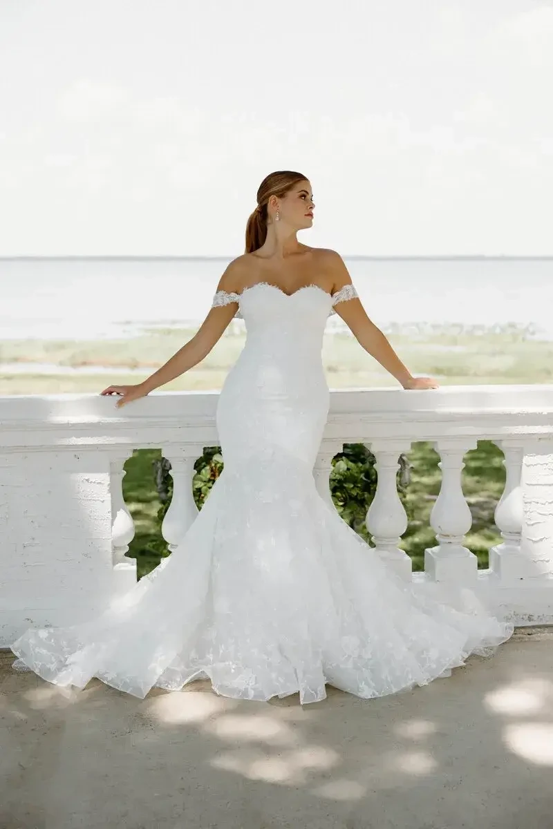 Bride in a lace off-the-shoulder gown stands gracefully on a balcony, overlooking a serene lake. The scene conveys elegance and tranquility.